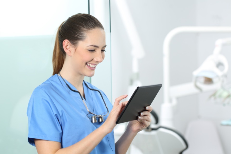 Female dentist wearing scrubs standing in dental room with glasses around neck holding a tablet and smiling