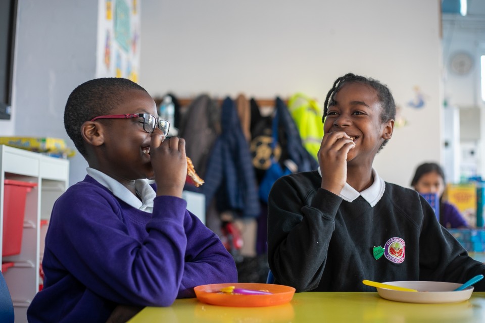 Two school children in uniform eating toast and laughing in classroom