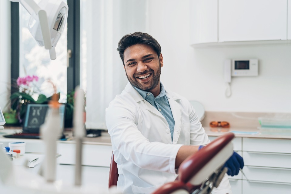 Male dentist wearing white coat and gloves sitting in dental room leaning on chair and laughing