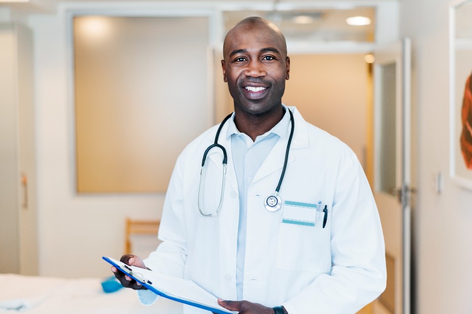 Male doctor smiling standing in corridor wearing white coat and stethoscope holding clipboard