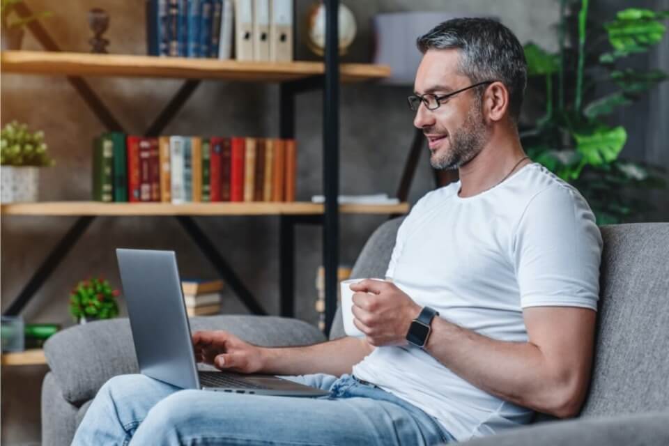 Man relaxing on sofa with laptop resting on laps while holding a mug