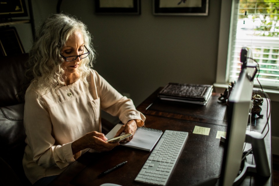 Professional woman sitting at desk in home office wearing glasses and using her phone
