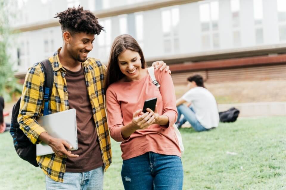 Male and female student standing together outside both looking at a phone smiling
