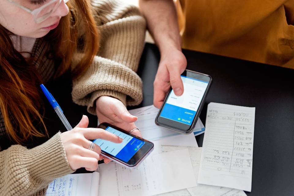 Two students using banking app on phones with bill papers on table