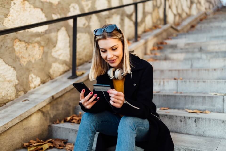 Young woman sitting outside on steps holding phone and bank card in hands