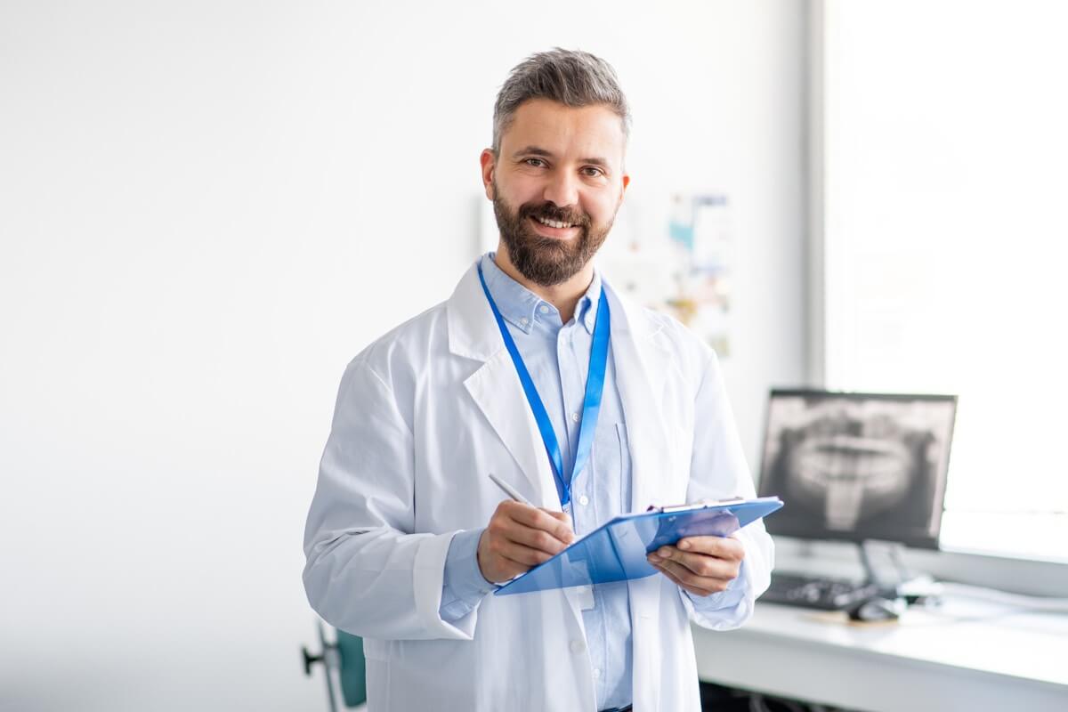 Male dentist in dental room wearing a white coat and holding a clipboard Male dentist in dental room wearing a white coat and holding a clipboard