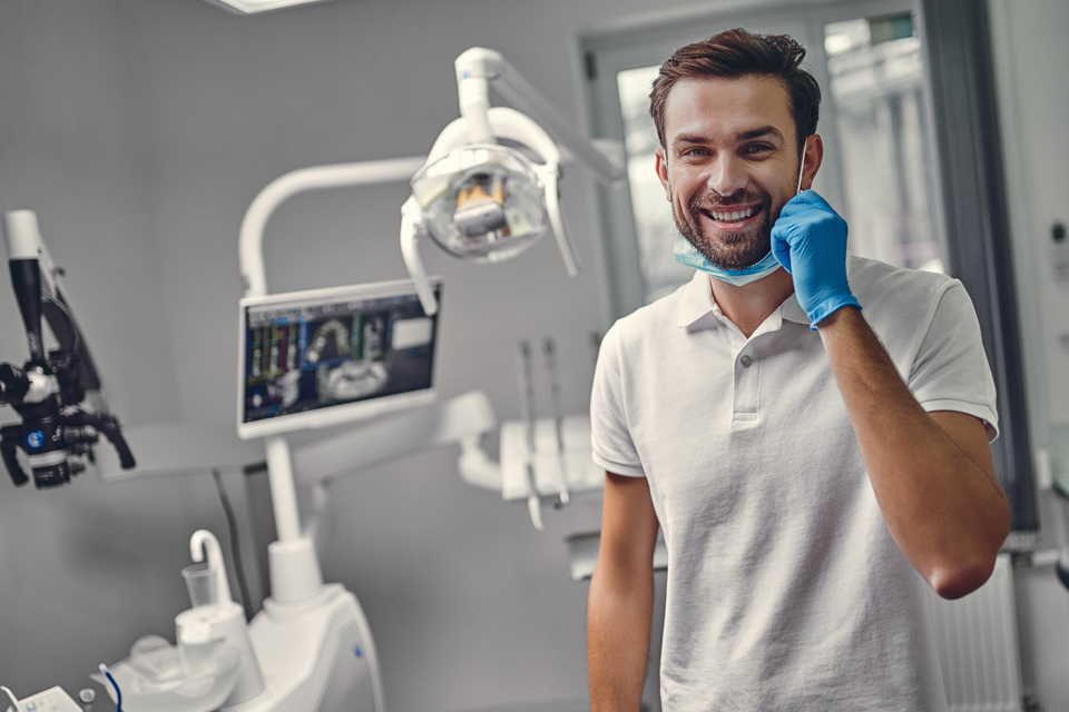 Young male dentist in PPE pulling down mask to smile