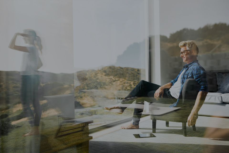 Mature man in chair looking through window at younger woman looking at the landscape