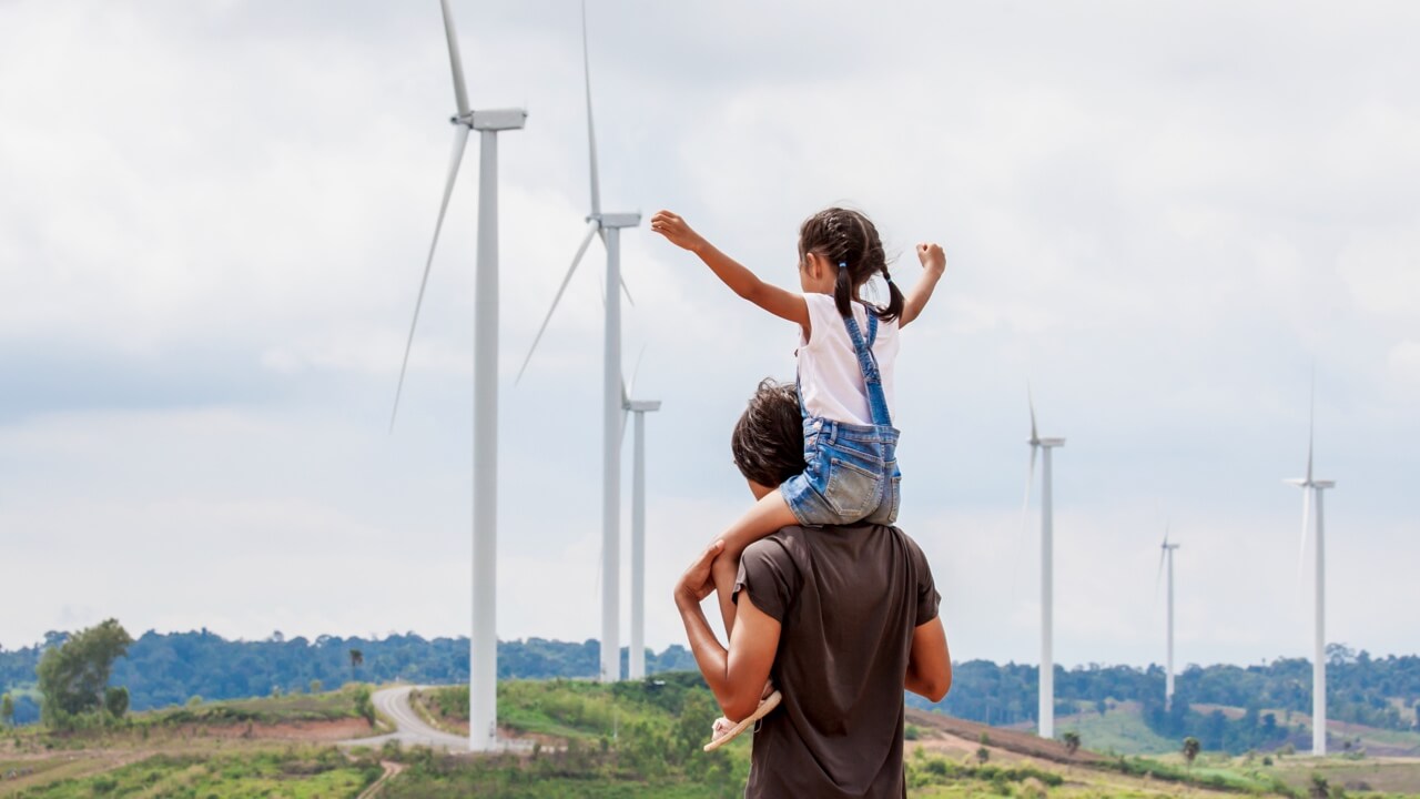 Young child sits on mans shoulders with arms open in front of wind turbines