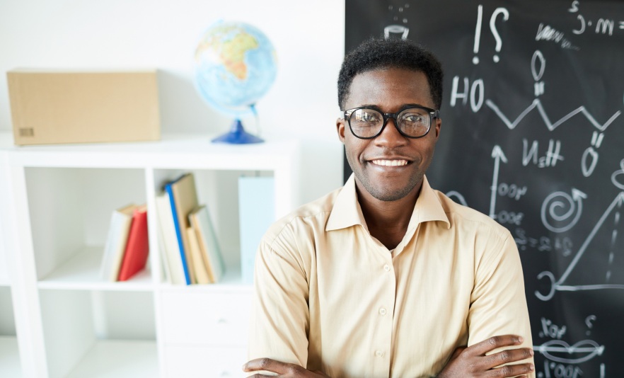 Male teacher standing in front of blackboard and bookshelves in classroom