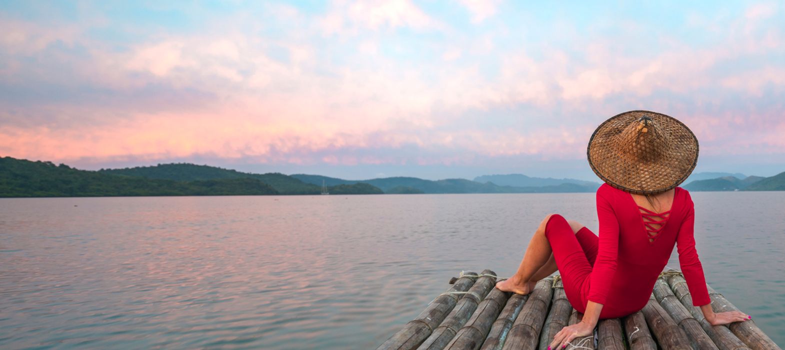 Woman sitting on wooden pier wearing a sunhat and looking at sunset