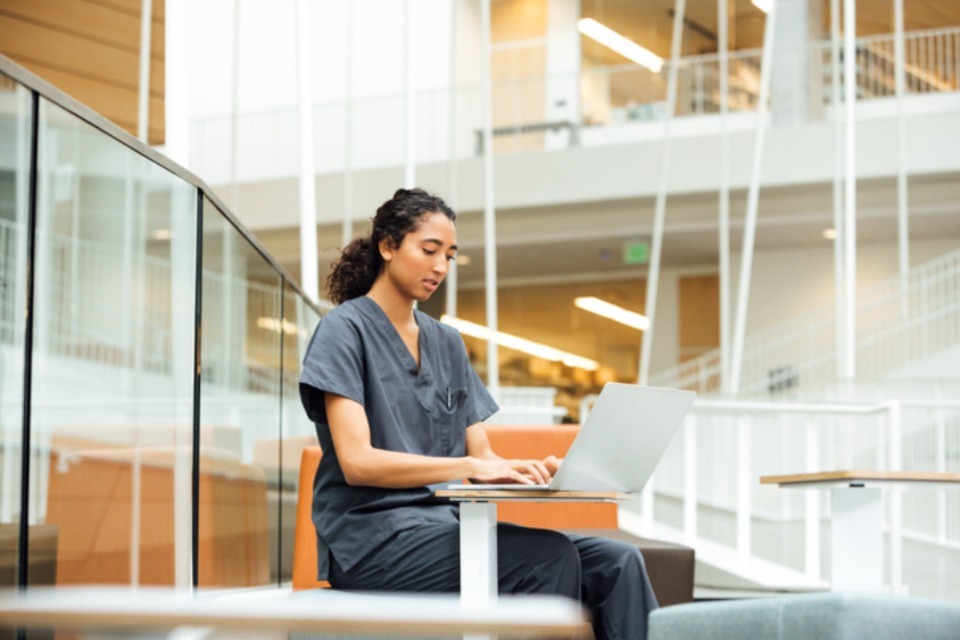 Female student sitting at table with laptop wearing scrubs